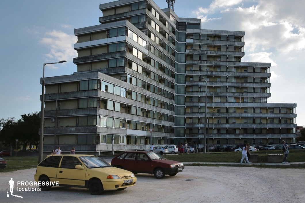 The image features a strikingly angular, tiered residential complex with a brutalist architectural style, characterized by its raw concrete facade, repetitive glass-paneled windows, and linear balcony structures. The building's staggered form creates a dynamic visual rhythm, while its weathered surfaces add a sense of age and authenticity. The open space in the foreground, with sparse greenery and an unpaved lot, enhances the starkness of the structure and provides a utilitarian, almost desolate atmosphere. This location is well-suited for dystopian narratives, social realism films, or crime dramas, offering a gritty, post-Soviet-era aesthetic that can evoke themes of isolation, urban decay, or societal tension.