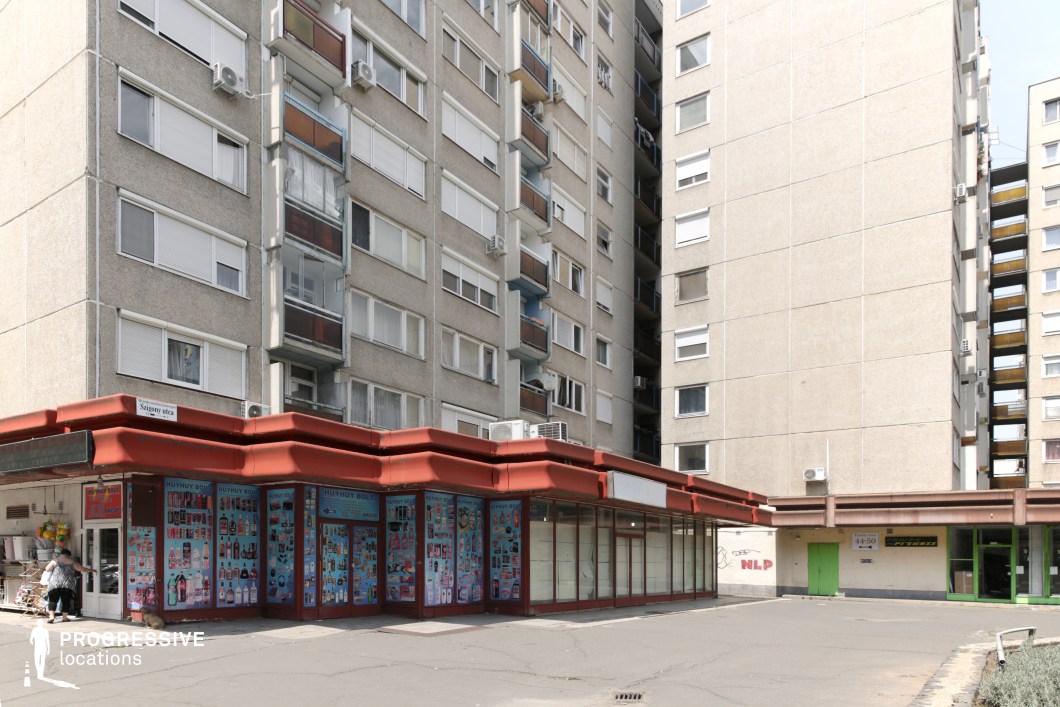 This urban setting features a towering brutalist-style apartment block with repetitive balconies and air conditioning units, creating a dense, functionalist aesthetic. At the base of the residential structure, a single-story commercial strip with a distinctive red overhang adds contrast to the otherwise gray concrete surroundings. Some storefronts appear active, while others are vacant or repurposed, contributing to a layered visual of economic transition. The wide, open pavement and lack of ornamentation enhance the stark, practical nature of the environment. This location is well-suited for films exploring themes of urban survival, social disparity, and everyday life in post-socialist or working-class communities, making it ideal for dramas, crime stories, or gritty realism.