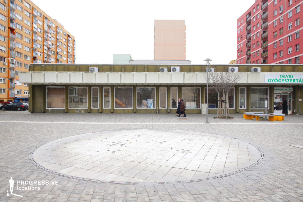 This urban scene captures a small commercial plaza with a low-rise, brutalist-style building featuring a repetitive window pattern, barred entrances, and an overhanging roof. The facade, weathered with greenish paneling and fitted with multiple air conditioning units, contrasts with the vibrant high-rise residential buildings in the background, painted in warm orange, red, and pastel hues. The foreground showcases a paved public square with a circular design element, adding a structured focal point to the space. The juxtaposition of aging retail infrastructure with modernized housing blocks makes this an ideal location for films exploring themes of urban transformation, working-class life, or gentrification. This setting is particularly well-suited for social dramas, crime stories, or narratives focusing on the intersection of past and present in post-socialist environments.