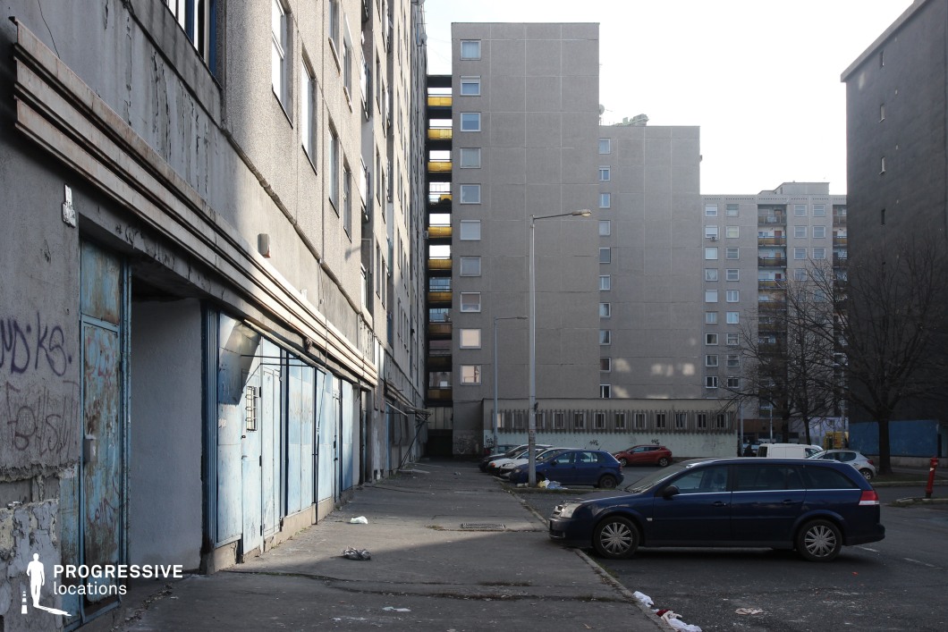 The image captures a stark, utilitarian residential area framed by aging concrete apartment buildings with minimal ornamentation. The narrow alleyway between the structures emphasizes a sense of confinement, while the worn facades, exposed stairwells, and graffiti-marked surfaces add an element of urban decay. The blue metal doors on the left, possibly for storage or maintenance spaces, show signs of rust and wear, reinforcing the gritty atmosphere. The scene is defined by muted colors, rigid geometric forms, and strong vertical lines, creating a visually raw and industrial aesthetic. This location is well-suited for crime dramas, social realism films, or dystopian settings, offering an evocative backdrop for narratives of isolation, survival, or urban tension.