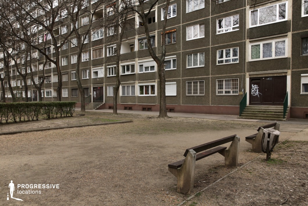 The image captures a stark residential courtyard in front of a mid-century panel-style apartment building with a textured, dark green facade. The repetitive pattern of windows and balconies creates a uniform, utilitarian aesthetic, while leafless trees and sparse hedges break the rigid geometry. In the foreground, worn concrete benches with wooden seating face an open dirt-covered communal area, contributing to an atmosphere of aging infrastructure and minimal upkeep. The graffiti-marked entrance and metal-barred windows reinforce the sense of urban grit. This location is ideal for social realism films, crime dramas, or psychological thrillers, providing a raw, lived-in backdrop that conveys themes of isolation, nostalgia, or working-class urban life.
