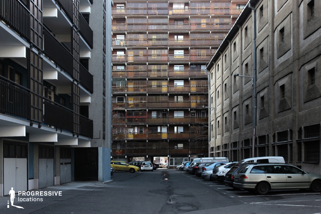 The image presents a narrow urban alleyway enclosed by three distinct architectural styles. On the left, a modern residential building features dark metal railings and recessed balconies, creating a rigid geometric pattern. On the right, a stark industrial-style structure with a rough concrete facade and repetitive square windows adds a sense of history and functionality. The background is dominated by a tall, aging residential block with a grid-like arrangement of balconies enclosed by rust-colored metal railings, giving the scene a worn, utilitarian aesthetic. The alley's confined space and contrasting materials create a visually intense atmosphere, making this location ideal for filming gritty crime dramas, neo-noir thrillers, or dystopian urban settings where tension and isolation play a key role.