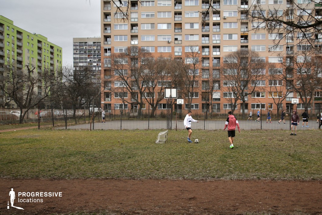 The image captures an urban sports field set against a backdrop of mid-rise apartment buildings with muted earth-toned facades. The field consists of a fenced basketball court and an adjacent grassy area with a small, weathered soccer goal. Surrounding the space, leafless trees create a visual contrast between the natural and built environment, while the worn fence and uneven grass hint at an aging communal area. The apartment buildings in the background display a mix of window styles and balconies, adding to the layered, lived-in aesthetic of the setting. This location is ideal for coming-of-age films, urban dramas, or sports-themed narratives, offering a raw, authentic atmosphere that reflects everyday community life.