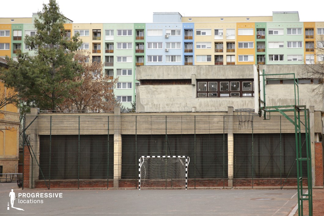 This urban sports court is enclosed by a tall, metal wire fence, reinforcing a sense of containment within the space. The court features a worn goalpost and remnants of a basketball hoop, set against a stark, brutalist-style concrete structure with small, broken windows, suggesting abandonment or neglect. Beyond this, a row of pastel-colored apartment buildings in shades of blue, green, and yellow contrasts sharply with the raw industrial appearance of the foreground. The juxtaposition of harsh, institutional design with lively residential facades creates a layered, visually complex setting. This location is well-suited for gritty dramas, social realism, and coming-of-age stories, especially those focusing on youth culture, urban decay, or economic disparity.