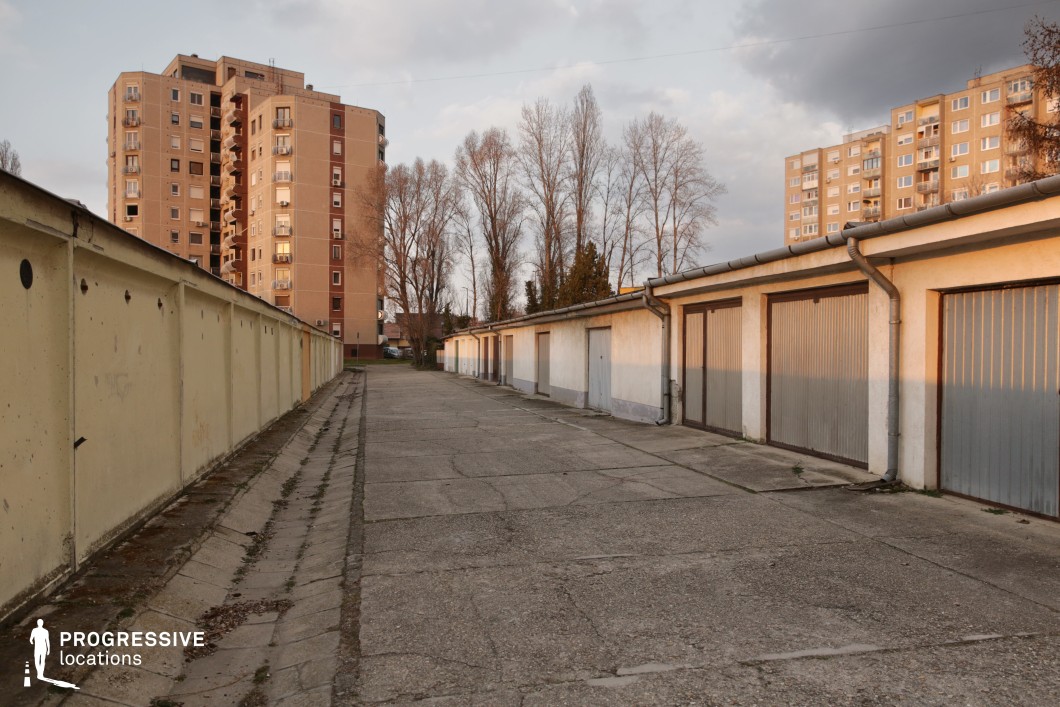 This urban location features a long, narrow alley lined with single-story garages on one side and a tall, weathered concrete wall on the other. The cracked pavement and aged facades suggest decades of use, while rusting metal doors and exposed drainage infrastructure add to the sense of decay. In the background, towering apartment blocks rise above the garages, reinforcing the contrast between small-scale, personal storage spaces and large-scale residential structures. The alley’s linear perspective creates depth, drawing attention toward the distant vanishing point. This setting is ideal for crime dramas, neo-noir thrillers, or urban suspense films, providing an atmospheric backdrop for clandestine meetings, escape sequences, or scenes of solitude and tension.