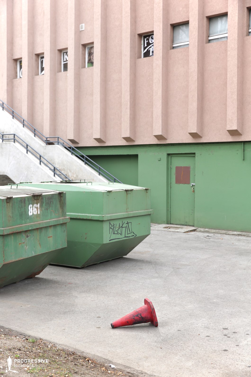 This urban setting features the rear side of a brutalist or late-modernist building, characterized by its rigid vertical ridges and utilitarian design. The structure’s lower section is painted in muted green, with a closed, worn-out metal door reinforcing a sense of restriction and disuse. A set of concrete stairs with metal railings leads to an upper level, adding depth and elevation to the scene. In the foreground, industrial waste containers and a cracked pavement contribute to the raw, unpolished atmosphere. The location’s harsh, functionalist aesthetic makes it an ideal setting for gritty crime dramas, neo-noir thrillers, or urban dystopian narratives exploring themes of abandonment, secrecy, or hidden activity within the cityscape.