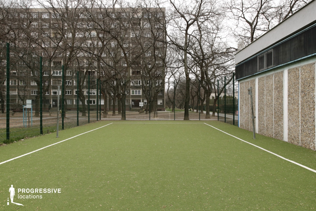 The image features a small artificial turf sports field enclosed by a tall metal fence, set within a residential neighborhood. The background consists of a mid-century apartment block with a repetitive facade, partially obscured by leafless trees, creating a contrast between the built environment and natural elements. To the right, a low-rise structure with a textured stone-like exterior adds another layer of urban functionality. The orderly lines of the sports field contrast with the more organic, slightly overgrown surroundings, emphasizing the structured yet aged nature of the space. This location is ideal for urban dramas, coming-of-age films, or sports-related narratives, offering a raw, authentic backdrop that blends community life with an underlying sense of nostalgia and quiet isolation.