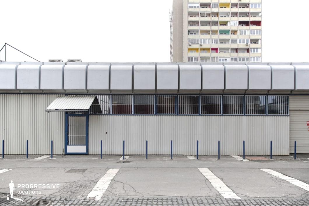 This location presents a stark contrast between a low-rise industrial or commercial warehouse and a towering residential block in the background. The warehouse features a utilitarian design with corrugated metal siding, barred windows, and a rounded overhanging roof, reinforcing an institutional, almost militaristic aesthetic. The entrance, marked by a small awning and a locked blue door, emphasizes security and restricted access. In the foreground, an empty asphalt lot with faded pedestrian crossings and cracks in the pavement enhances the atmosphere of neglect and urban wear. The rigid, repetitive balconies of the apartment complex in the background provide a visual counterpoint, reinforcing themes of division and urban decay. This location is ideal for crime thrillers, dystopian narratives, and social dramas exploring themes of isolation, security, and economic decline.