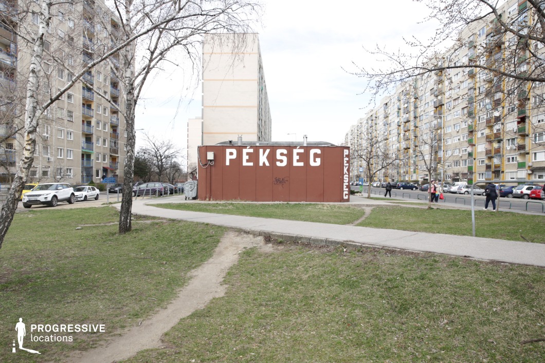 This urban setting showcases a compact, standalone bakery with a brown metal facade, positioned between towering panel-style apartment buildings. The structure appears temporary or prefabricated. The surrounding area consists of a mix of paved walkways and informal dirt paths cutting through patches of grass, suggesting frequent pedestrian movement. The background is dominated by rows of dense, uniform residential blocks with repetitive balconies and windows, emphasizing the mass-housing aesthetic. This location is ideal for films exploring themes of working-class life, urban resilience, or local commerce, making it suitable for social dramas, slice-of-life narratives, or neo-realist storytelling.