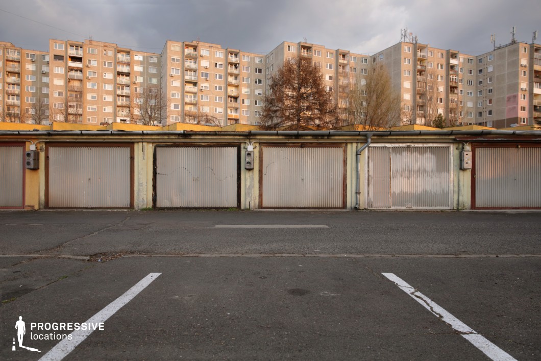 The image showcases a row of aged, metal garage doors with faded yellow and rust-streaked facades, positioned against a backdrop of uniform, Soviet-style apartment blocks. The garages, with their worn-out surfaces and utilitarian design, contrast sharply with the towering residential buildings above, creating a layered urban environment. The foreground features an asphalt parking lot with faded white lines and visible cracks, enhancing the gritty, lived-in aesthetic of the setting. The combination of decayed infrastructure and repetitive architectural patterns makes this location a strong visual choice for crime thrillers, neo-noir films, or urban dramas exploring themes of isolation, secrecy, or working-class struggles.