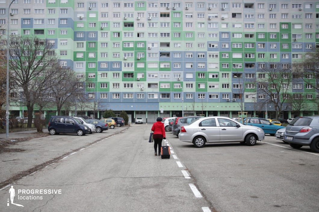 This urban setting features a towering panel-style apartment block with a gradient facade of blue and green, adding a distinct visual identity to the otherwise uniform architecture. The foreground consists of an expansive, weathered parking lot with faded white markings and a variety of parked vehicles. The linear perspective leads directly to the residential building’s ground-level passage, creating a sense of depth and symmetry. The repetitive windows and balconies emphasize the dense, structured nature of the housing estate, evoking themes of collectivism and urban routine. This location is ideal for social realism, coming-of-age stories, and crime dramas, offering an authentic backdrop for narratives exploring everyday life, migration, or the contrasts between personal and communal existence.