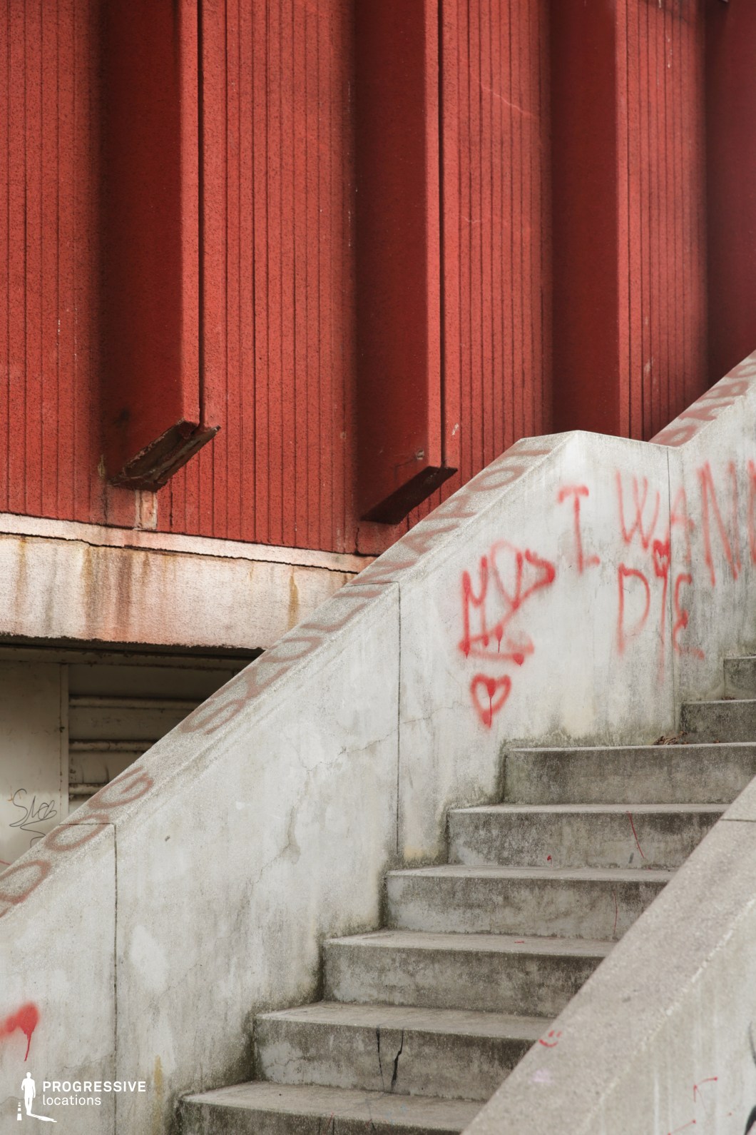This urban setting features a worn concrete staircase leading up against a striking red industrial-style facade with vertical paneling. The stark geometry of the scene is softened by graffiti markings on the stair rail, adding a layer of urban decay and social commentary. The red exterior panels, accented by rust and peeling paint, contrast with the cold, aged concrete steps that show visible cracks and discoloration. The overall atmosphere is one of neglect and raw authenticity, making this an ideal location for films exploring urban alienation, underground culture, or gritty realism. This setting is particularly well-suited for crime dramas, psychological thrillers, and social commentaries focused on inner-city life.