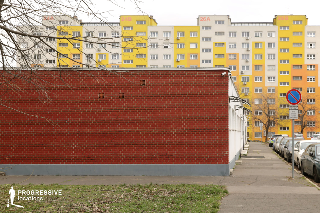 This urban scene presents a strong contrast between the foreground and background, with a bold red brick wall dominating the lower half of the image, while a bright yellow and beige high-rise housing block rises above it. The rigid, utilitarian architecture of the panel housing estate, complete with repetitive windows and balconies, contrasts with the industrial character of the low-rise building. The street beside the wall, with a pedestrian sidewalk, adds an additional layer of urban realism. The symmetrical framing and strong color contrasts make this an ideal location for films exploring themes of social hierarchy, division, or modern urban life, particularly in crime thrillers, social dramas, or neo-noir settings.