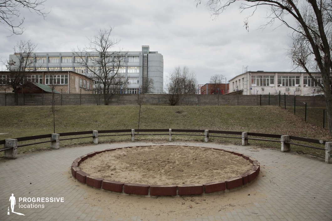 The image features a circular sandpit bordered by red bricks, set in a paved open area enclosed by aged concrete benches. A grassy incline separates the foreground from the background, where a tall concrete wall, lined with graffiti, encloses an institutional complex composed of industrial-style buildings with a mix of brick, metal, and glass facades. The rigid geometry of the structures contrasts with the organic shapes of leafless trees, adding depth to the composition. The muted color palette and minimalistic layout evoke a utilitarian, slightly desolate atmosphere. This location is well-suited for social realism films, institutional dramas, or psychological thrillers, offering a setting that conveys themes of isolation, nostalgia, or the passage of time.