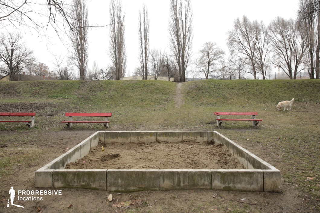 A quiet, open park space with a weathered concrete-bordered sandpit as its focal point, flanked by two red benches. The gently sloping grassy hill in the background, lined with tall, bare trees, adds depth and a natural barrier. A narrow dirt path leads over the hill, suggesting movement and continuity beyond the frame. The muted color palette and sparse elements create a setting that feels both tranquil and slightly melancholic, making it ideal 