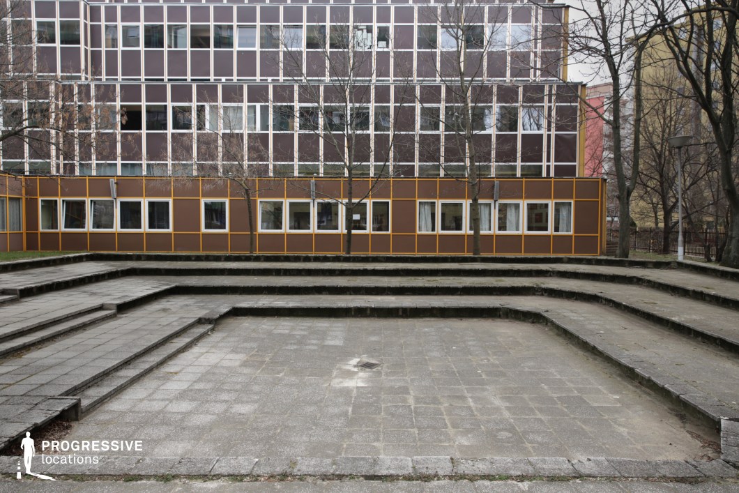 The image showcases a structured, tiered concrete courtyard leading up to a modular, mid-century modern school building with a grid-like facade. The building’s brown and yellow paneling, accented with white-framed windows, creates a repetitive, rigid aesthetic. The stepped courtyard, possibly designed as an outdoor gathering or performance space, appears aged with worn paving and overgrown edges, emphasizing a sense of neglect. Leafless trees line the perimeter, adding vertical contrast to the horizontal geometry of the architecture. The setting exudes an institutional atmosphere, making it ideal for filming school-based dramas, social realism narratives, or dystopian thrillers, with its raw, functional design enhancing themes of authority, nostalgia, or urban isolation.