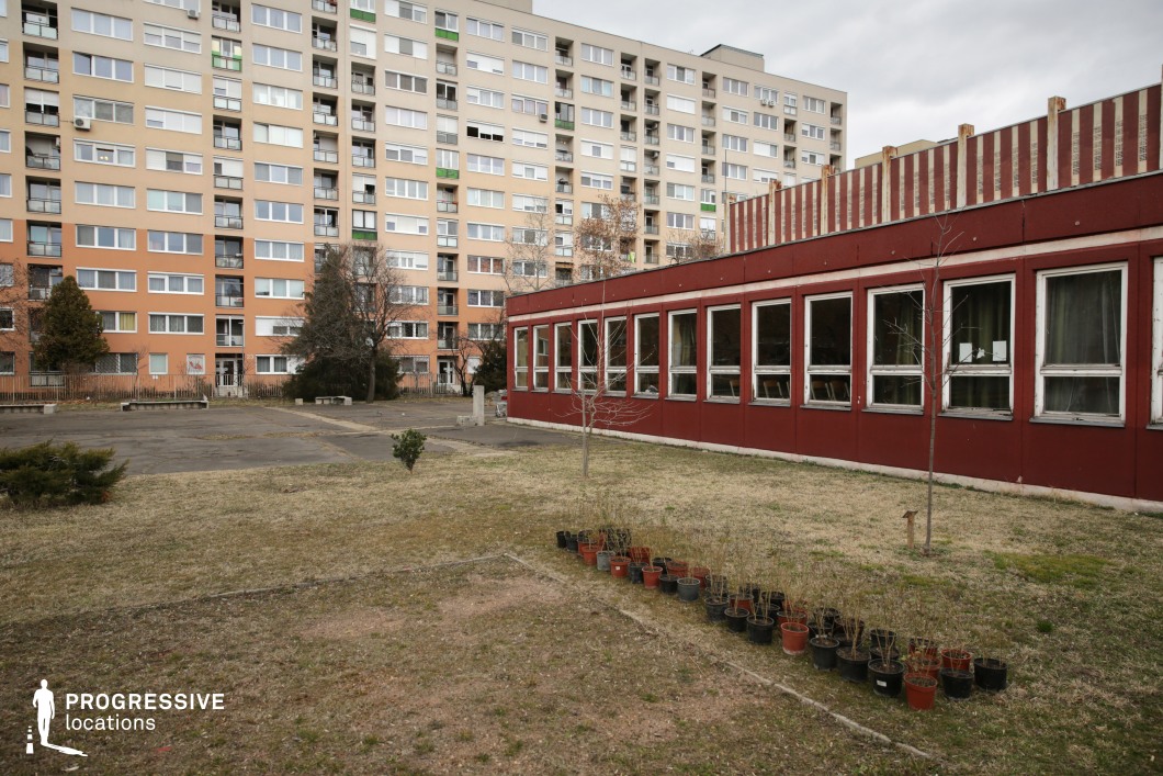 The image depicts a juxtaposition of urban residential and institutional architecture, featuring a large, multi-story apartment complex in the background and a single-story red-panel building in the foreground. The apartment block is characterized by repetitive windows and balconies, with a neutral-toned facade accented by orange and beige hues, reflecting a functionalist or socialist-era architectural style. The foreground structure, likely a school or community center, has a modular design with a red exterior and a row of rectangular windows, some of which are partially open or show signs of wear. The surrounding outdoor space consists of a patchy grassy area with a row of potted plants and a paved section, possibly a former courtyard or playground. The slightly weathered look and utilitarian layout of the location create an ideal backdrop for films requiring an authentic, raw urban environment, particularly in social dramas, crime narratives, or stories exploring post-Soviet or Eastern European aesthetics.