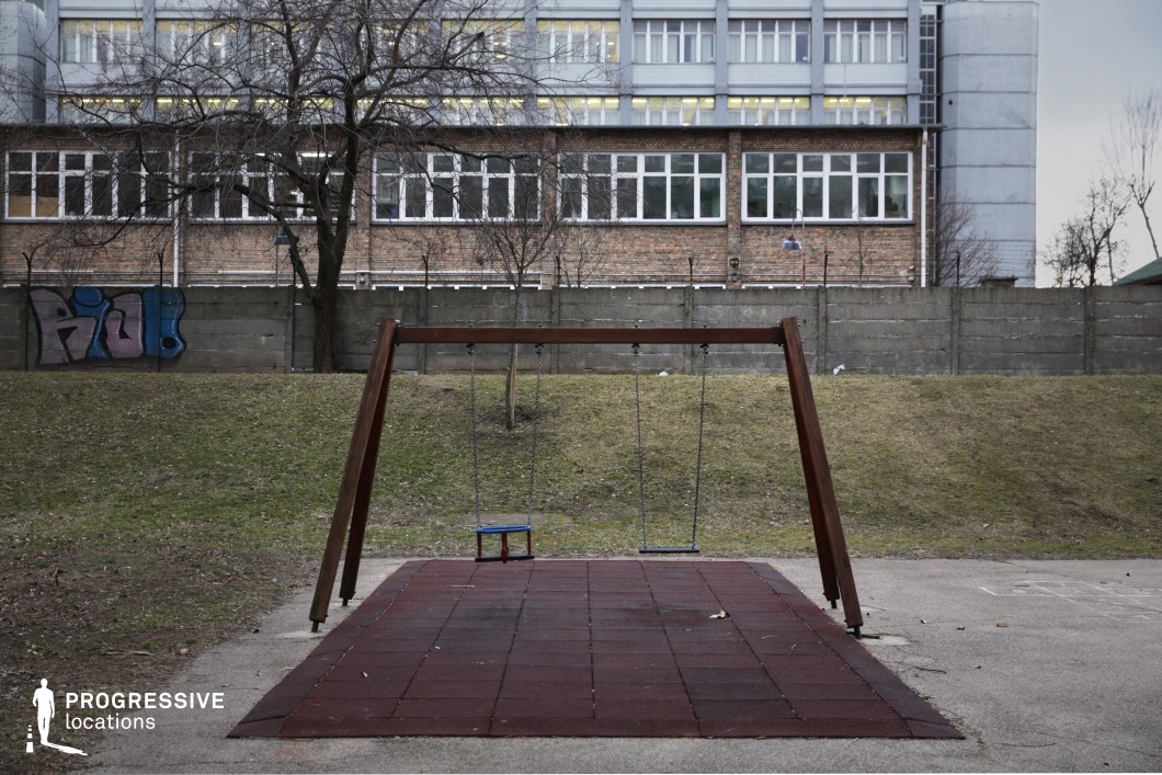 The image presents a solitary wooden swing set with worn chains, positioned on a rubber mat in a sparse urban playground. The surrounding area consists of a grassy incline leading up to a tall concrete wall topped with barbed wire, separating the playground from an industrial-style brick and metal building with rows of windows. Graffiti on the wall and the utilitarian architecture contribute to a raw, unembellished atmosphere. The contrast between the playground equipment and the rigid, institutional background creates a visually striking and emotionally evocative scene. This location is ideal for social realism films, psychological dramas, or dystopian settings, offering a backdrop that conveys themes of isolation, nostalgia, or urban decay.