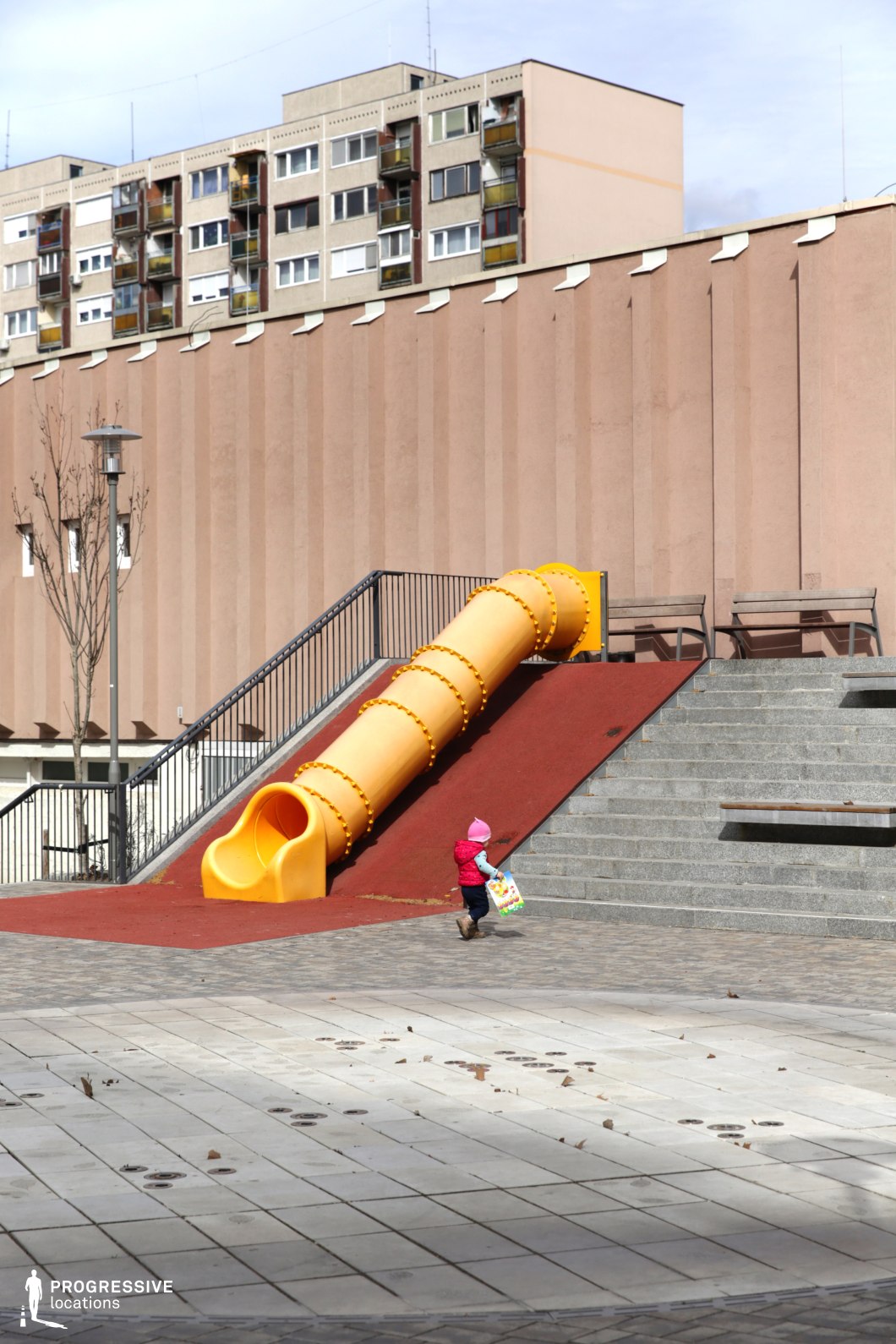 This urban playground is set against a stark, monolithic concrete structure with vertical ridges, evoking a brutalist or late-modernist architectural style. A bright yellow enclosed slide descends from an elevated platform, contrasting sharply with the muted tones of the surrounding environment. Wide concrete steps and built-in benches suggest a multi-functional public space, blending recreation with structured urban design. In the background, a row of panel-style apartment buildings with repetitive balconies and windows reinforces the dense, residential setting. The juxtaposition of playful elements against rigid, imposing architecture makes this location ideal for films exploring themes of childhood in an urban landscape, nostalgia, or the contrast between personal freedom and institutional control, fitting for social dramas, dystopian settings, or coming-of-age narratives.