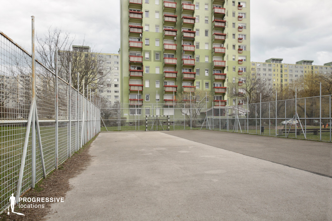 This urban sports court is surrounded by a tall metal fence, creating a confined yet functional recreational space. The concrete and asphalt playing surface is well-worn, indicating frequent use, while a small goalpost at the far end suggests its primary function for casual football games. The background is dominated by a large, uniform apartment block with a green facade and red balcony railings, characteristic of post-socialist residential architecture. The repeating balconies and rigid structure contrast with the open court, reinforcing the visual tension between restriction and play. This setting is ideal for films exploring themes of youth culture, social realism, or urban struggles, making it well-suited for coming-of-age stories, sports dramas, or narratives centered on working-class communities.