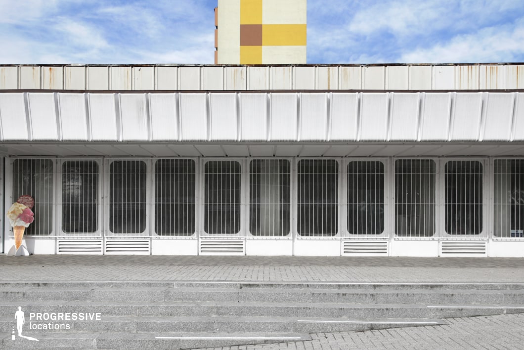 This imposing urban structure features a stark, repetitive facade with tall, barred windows and a corrugated overhang, emphasizing a brutalist or late-modernist architectural style. The white exterior is slightly weathered, with visible rust on the edges of the metal panels, reinforcing an atmosphere of urban decline or underutilization. The foreground consists of wide, symmetrical concrete stairs leading to the entrance, adding a sense of formality and rigidity to the composition. A fragment of a colorful high-rise apartment block in the background contrasts with the monochrome structure, subtly hinting at the surrounding residential environment. This setting is ideal for dystopian films, crime thrillers, or social dramas, evoking themes of isolation, institutional control, or economic stagnation in a post-socialist urban landscape.