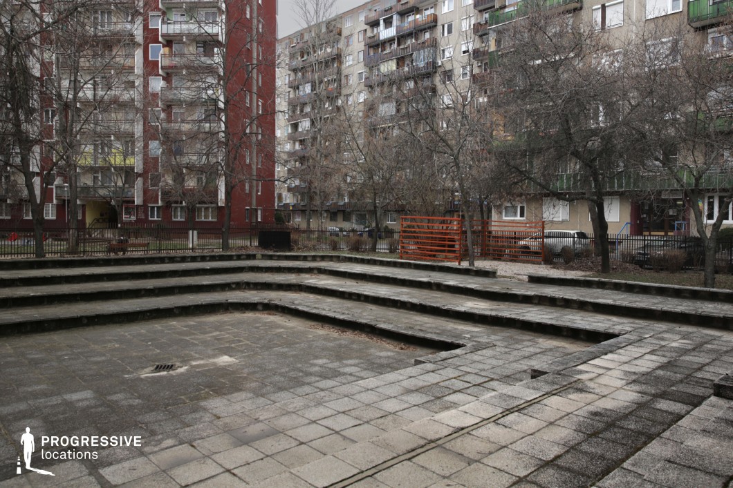 The image showcases a weathered, tiered concrete amphitheater set within a residential courtyard, surrounded by mid-century apartment buildings with muted facades and repetitive balconies. The open, geometric structure of the stepped plaza provides a sense of symmetry, while the worn surfaces, cracks, and scattered debris contribute to an aged, utilitarian atmosphere. Leafless trees and a rust-colored metal fence enclose the space, adding a sense of separation from the surrounding urban landscape. The contrast between the rigid built environment and the organic elements enhances the stark, functional aesthetic. This location is ideal for urban dramas, social realism films, or crime thrillers, offering a visually compelling setting that conveys themes of nostalgia, decay, or quiet tension.