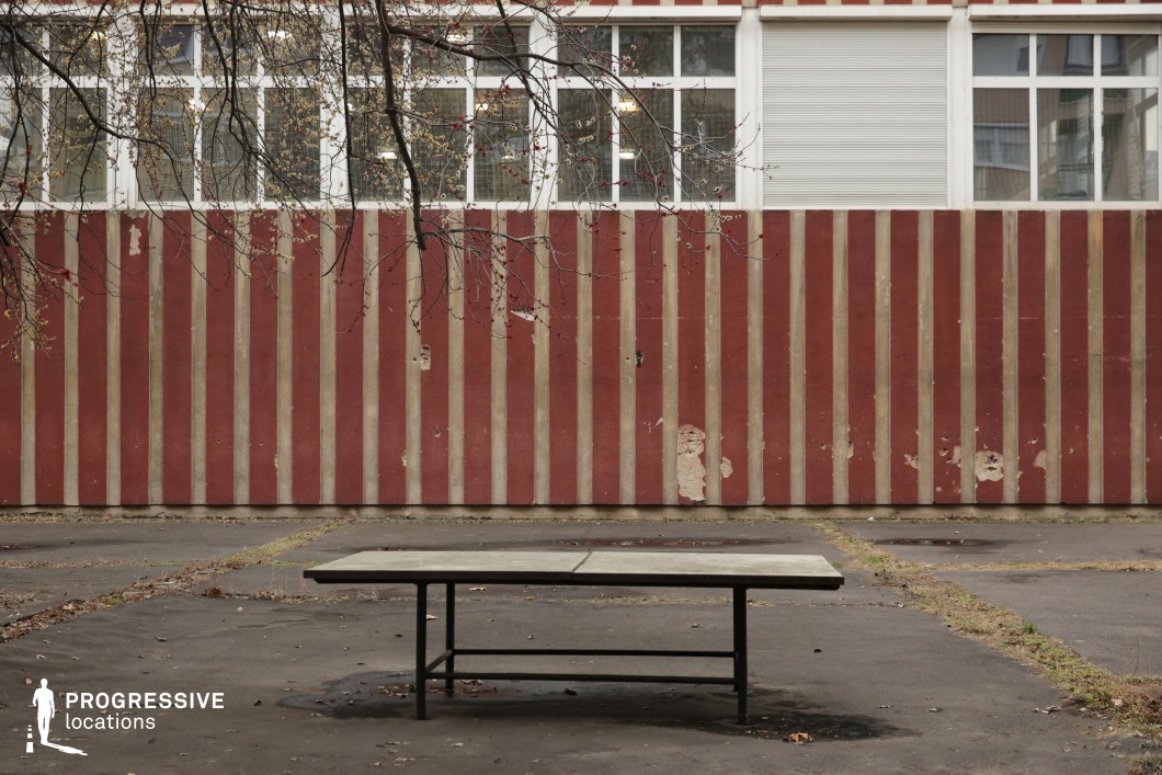The image captures an austere outdoor setting dominated by a weathered concrete table in the foreground, positioned on a worn asphalt surface. The backdrop features a red-and-beige striped institutional building with peeling paint and barred windows, suggesting a utilitarian or aging public facility. The symmetrical facade, reinforced by the rigid vertical pattern of the wall, emphasizes a sense of order and confinement. The stark contrast between the structured architecture and the abandoned, neglected atmosphere makes this location ideal for filming scenes that evoke isolation, nostalgia, or societal decay. It is well-suited for dramas, psychological thrillers, and dystopian narratives exploring themes of abandonment or institutional rigidity.