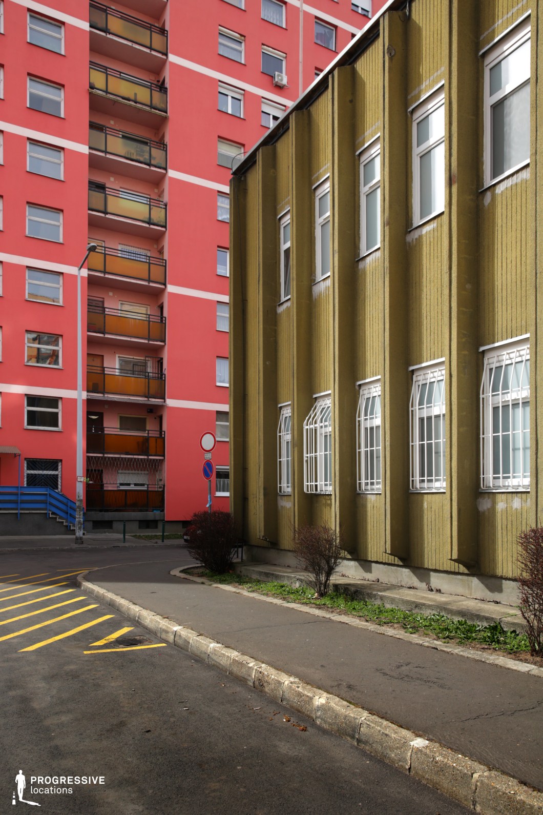 This urban location presents a striking visual contrast between two distinct architectural styles. On the left, a vibrant red high-rise residential building with uniform balconies and repetitive window patterns embodies contemporary housing renovations. On the right, an older institutional or commercial structure with a muted olive-green facade, vertical ridges, and barred windows exudes a more austere, utilitarian aesthetic. The juxtaposition of these structures, along with the layered urban elements such as pavement markings, signage, and fencing, creates a dynamic environment. This location is well-suited for films exploring themes of modernization versus decay, urban transitions, or socioeconomic contrasts, making it ideal for social dramas, crime thrillers, or neo-noir storytelling.