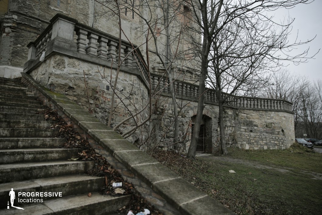 This image features an aged stone staircase, partially covered with fallen leaves, leading up to a fortress-like terrace with crumbling balustrades and arched brick openings. The walls, constructed from rough-hewn stone and weathered brick, display signs of decay, reinforcing the sense of abandonment. Leafless trees with twisted branches frame the structure, adding to the stark, desolate atmosphere. The muted color palette and overgrown surroundings enhance the setting’s eerie and historical character. This location is ideal for gothic thrillers, historical dramas, or mystery films, serving as a haunting backdrop for hidden pasts, secretive encounters, or explorations of forgotten places.