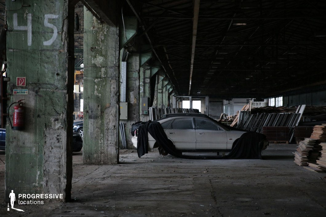 The image depicts a vast, decayed industrial warehouse with towering concrete pillars, peeling green paint, and a darkened ceiling supported by rusted metal beams. The worn concrete floor is scattered with debris, while stacks of disorganized metal frames and wooden planks line the space. A dust-covered car, partially draped in black fabric, sits eerily in the center, adding to the sense of abandonment. The vastness of the hall, combined with its dilapidated state, creates an atmospheric and cinematic setting. This location is ideal for crime dramas, neo-noir thrillers, or post-apocalyptic films, providing a raw and gritty backdrop for underground dealings, action sequences, or moments of suspense in forgotten industrial landscapes.