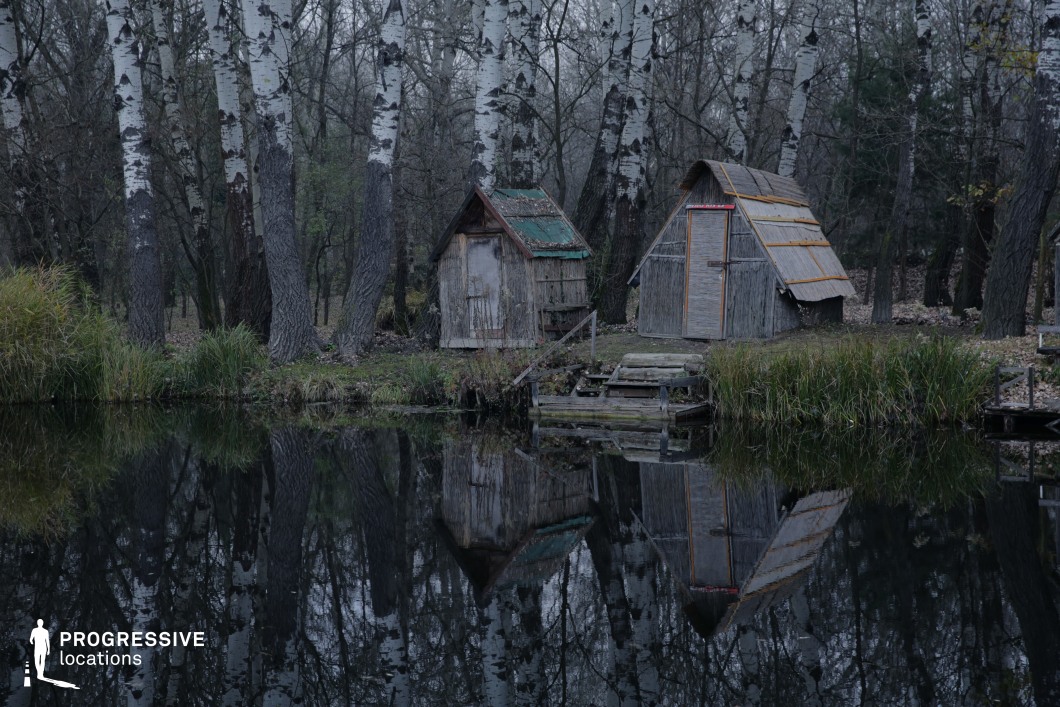 The image showcases two rustic wooden huts on the edge of a still lake, surrounded by tall birch trees with striking white bark. The structures appear weathered, with aged wooden planks and makeshift roofs, adding to the sense of remoteness. A small, decaying dock extends into the water, enhancing the abandoned feel of the setting. The reflection of the huts and trees in the water creates a symmetrical, almost surreal visual effect. This location is ideal for horror, mystery, or psychological thriller productions, offering an eerie, secluded atmosphere perfect for stories involving isolation, forgotten places, or unsettling discoveries.
