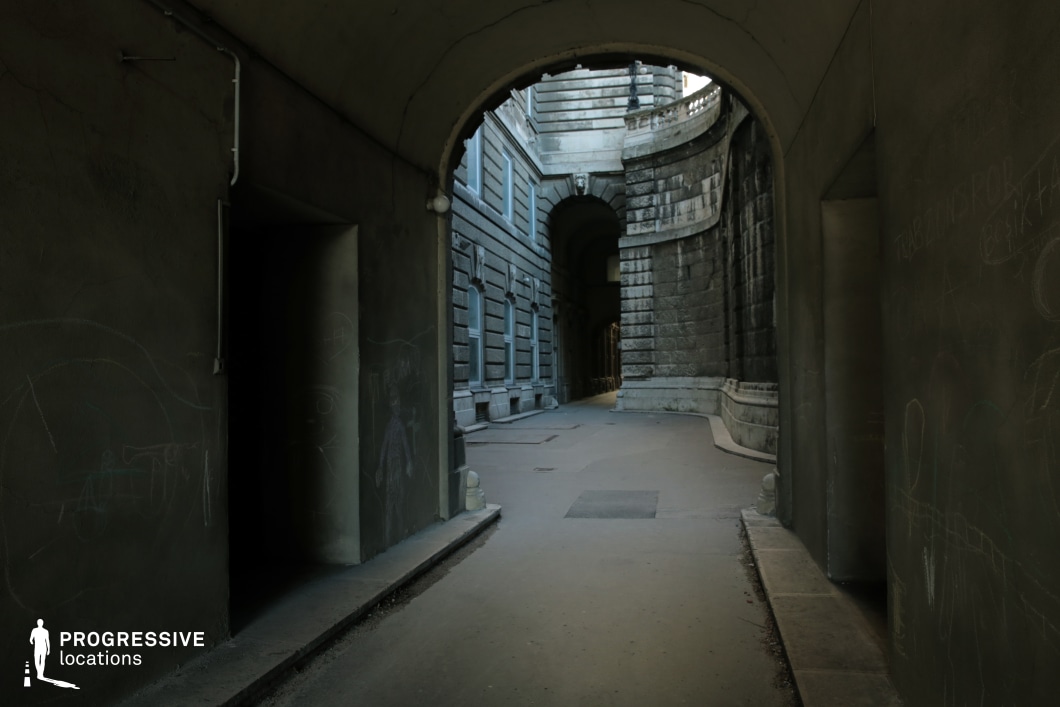 This secluded passageway, framed by a curved arch, leads through imposing stone architecture, creating a stark contrast between light and shadow. The weathered walls, adorned with subtle graffiti, enhance the location’s raw, urban character, while the worn pavement and narrow perspective add to the sense of depth. The heavy masonry and austere design evoke a historical or institutional atmosphere, making this an ideal setting for neo-noir thrillers, espionage narratives, or tense pursuit scenes. The interplay of light filtering through the passage adds a dramatic effect, heightening the sense of secrecy and hidden intrigue.