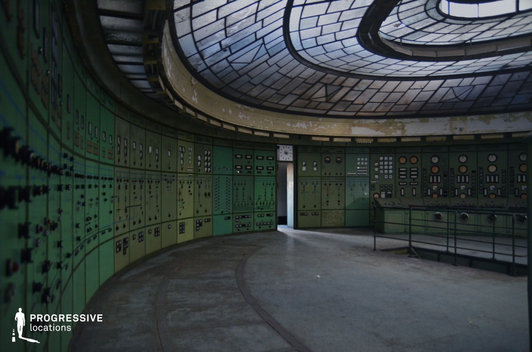 This abandoned control room features a striking curved layout with rows of aged green control panels covered in switches, dials, and analog gauges. The domed ceiling, adorned with a geometric glass skylight, filters soft, diffused light into the space, highlighting the peeling paint and worn surfaces. The room's symmetrical design and faded industrial aesthetic evoke a sense of lost technological grandeur. The setting is ideal for dystopian, sci-fi, or espionage-themed productions, offering a visually rich backdrop for tense decision-making sequences, secret operations, or retro-futuristic storytelling.