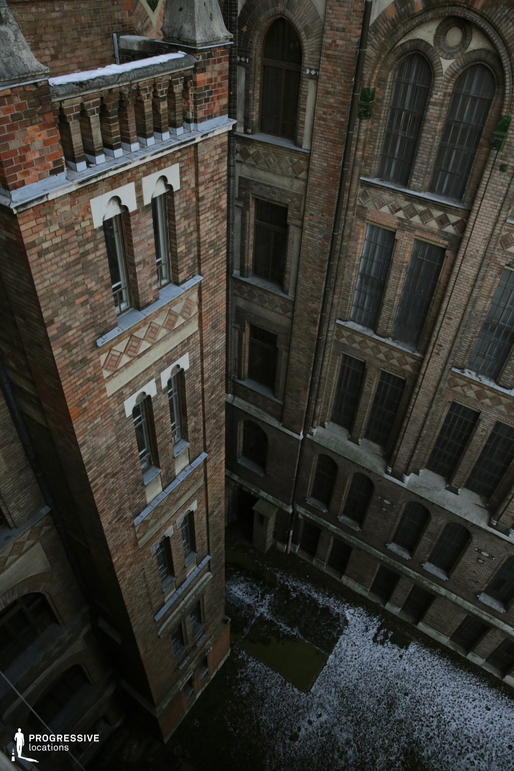 This image captures an enclosed courtyard surrounded by imposing red brick buildings with intricate detailing, arched windows, and decorative elements reminiscent of late 19th-century institutional architecture. The high vantage point emphasizes the verticality of the structures, while the shadowed lower courtyard, partially covered in patches of snow and moss, enhances the stark, abandoned atmosphere. The dark, closed windows add a sense of mystery and inaccessibility, reinforcing the setting’s ominous character. This location is ideal for psychological thrillers, historical dramas, or gothic horror films, providing a dramatic and confined backdrop for tense confrontations, hidden secrets, or eerie discoveries.
