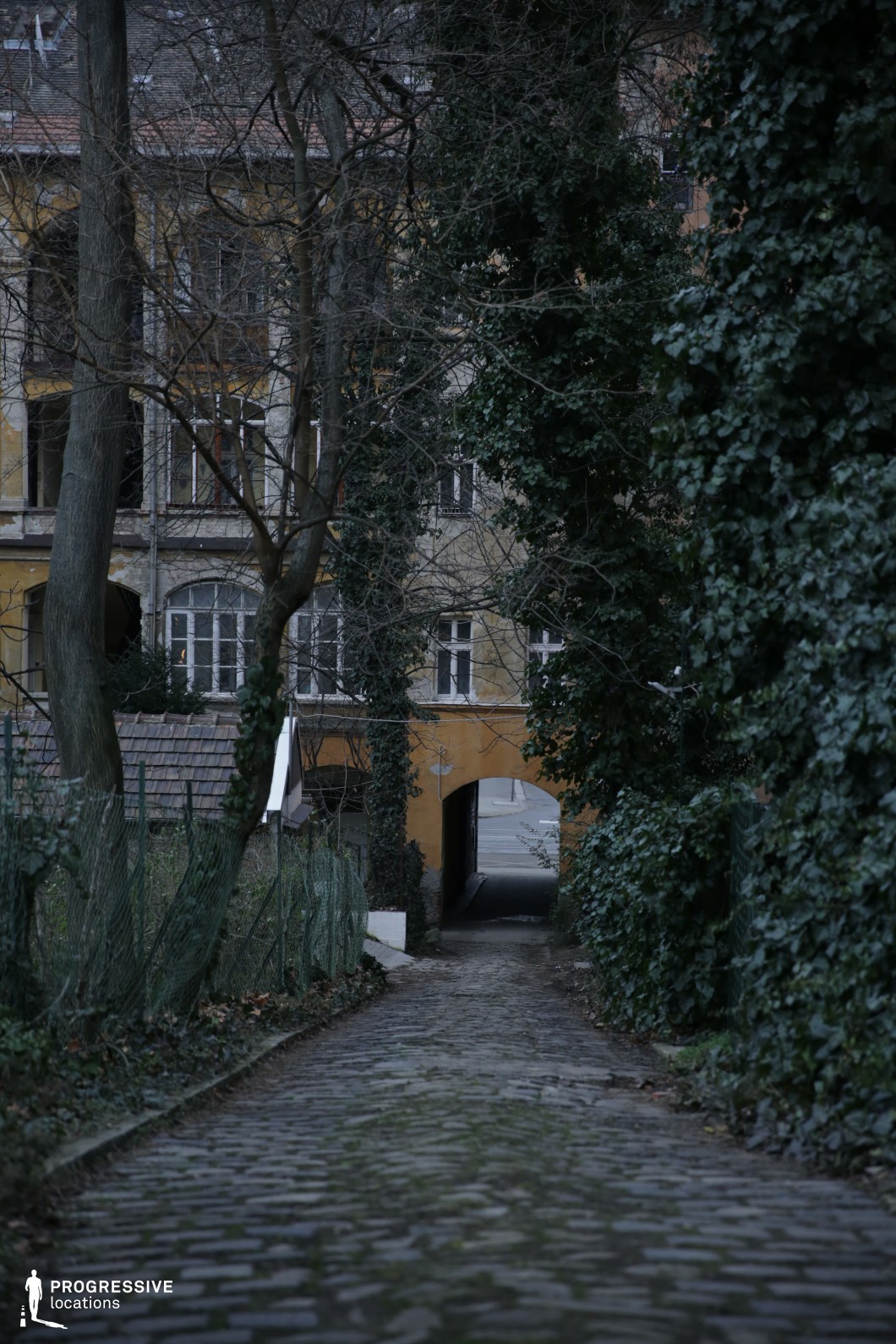 This image features a narrow, aged cobblestone path winding through an overgrown passage flanked by ivy-covered walls and barren trees. The pathway leads to a small, yellow-hued building with an arched passageway that opens onto a distant street, creating a sense of transition between hidden seclusion and urban life. The weathered facade, cracked fencing, and creeping vegetation enhance the scene’s aged, almost forgotten charm. This location is ideal for mystery thrillers, period dramas, or psychological narratives, offering a setting for moments of quiet introspection, secretive encounters, or eerie discoveries within a secluded urban space.
