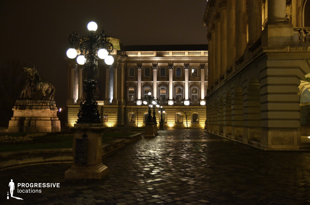 This stately courtyard is framed by imposing neoclassical buildings, accentuated by dramatic lighting that highlights their intricate facades, arched windows, and elegant columns. The polished cobblestone pathway reflects the glow of ornate street lamps, creating a striking contrast between the illuminated structures and the darkened surroundings. A commanding equestrian statue on a stone pedestal adds a sense of historical significance and grandeur to the scene. This location is ideal for period dramas, political thrillers, or mystery narratives, providing a regal yet enigmatic atmosphere suited for clandestine meetings, high-stakes confrontations, or moments of reflection in a historical setting.