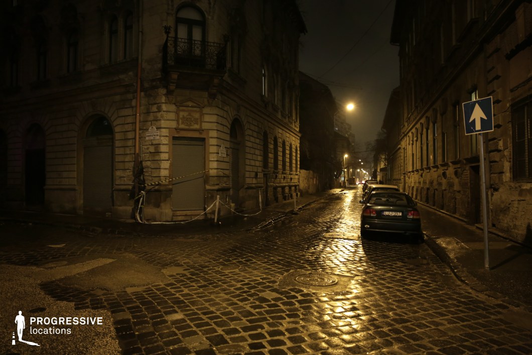 This image captures a dimly lit, narrow street flanked by historic, weathered buildings with intricate facades and arched windows. The cobblestone pavement, slick with moisture, reflects the warm glow of the streetlights, creating a dramatic interplay of light and shadow. A corner building with closed shutters and caution tape adds a sense of mystery, while the receding perspective of the street lined with parked cars enhances the depth and realism of the urban environment. The aged architecture and atmospheric lighting make this location ideal for noir films, crime dramas, and psychological thrillers, serving as a perfect setting for clandestine meetings, suspenseful pursuits, or introspective character moments.