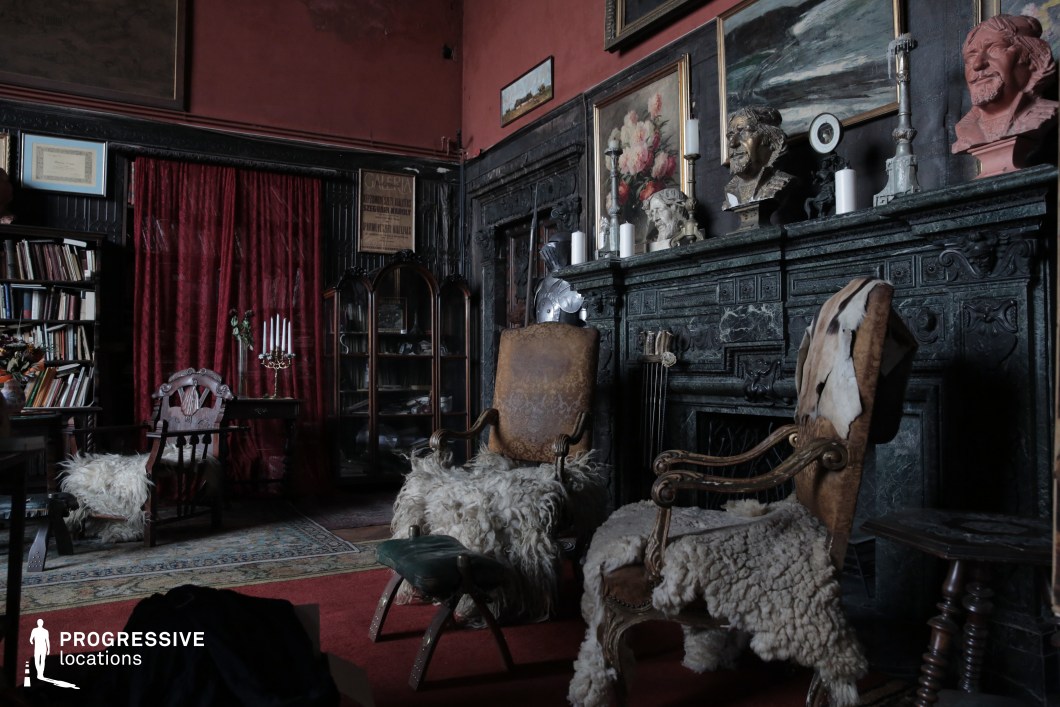 This richly decorated study exudes an air of mystery and old-world sophistication, with its dark green marble fireplace, heavy red drapery, and an eclectic mix of antique furniture and artwork. Ornate chairs draped in sheepskin, aged bookshelves, and classical busts contribute to the space’s intellectual yet slightly eerie ambiance. The dim lighting and deep color palette, combined with layered textures of wood, fur, and stone, create an environment that feels both luxurious and haunting. This location is ideal for gothic dramas, psychological thrillers, or period mysteries, serving as a compelling backdrop for scenes involving secretive discussions, introspective moments, or hidden intrigue within an aristocratic or academic setting.