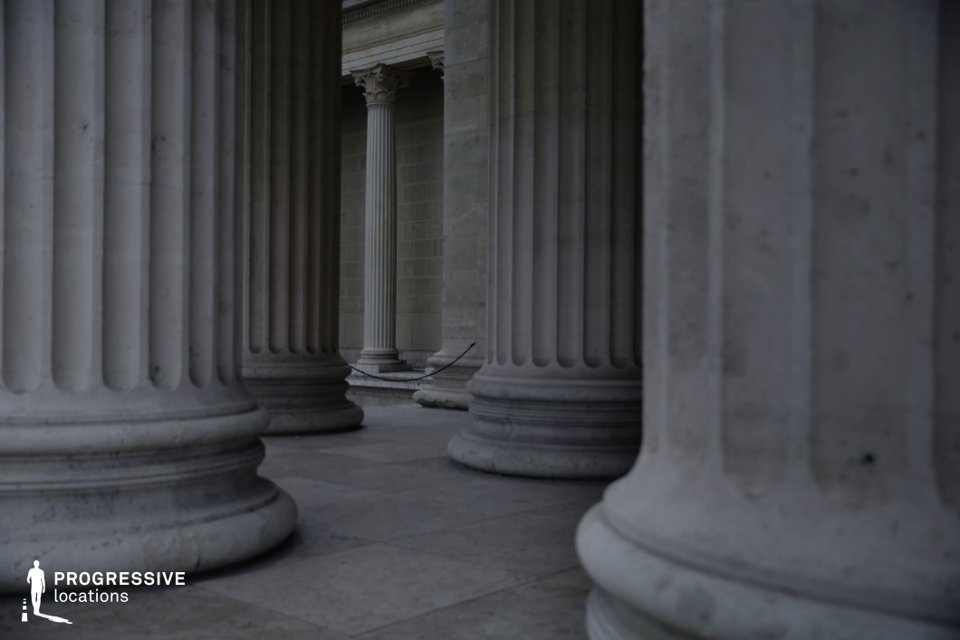 This image showcases a series of towering, fluted columns forming a grand, classical colonnade. The massive stone pillars, evenly spaced, create a strong sense of rhythm and depth, drawing the eye toward the vanishing point in the background. The soft natural light emphasizes the smooth surfaces and subtle imperfections in the stone, reinforcing the timeless elegance of the structure. The cool, muted tones enhance the imposing and almost austere atmosphere, making this location ideal for historical dramas, political thrillers, or philosophical narratives. The setting’s grandeur lends itself well to scenes involving power, judgment, or introspection, with the structured composition amplifying a sense of authority and permanence.