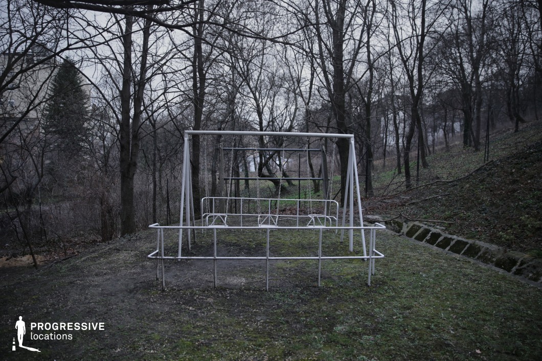 The image features a desolate metal playground structure standing alone in a wooded area, surrounded by leafless trees and overgrown grass. The stark white metal frame contrasts with the dark, natural tones of the environment, reinforcing a sense of abandonment and decay. The surrounding landscape slopes gently downward, with a distant path fading into the background, adding to the eerie isolation of the scene. This location is ideal for psychological thrillers, horror films, or dystopian dramas, providing a haunting backdrop for themes of lost innocence, forgotten spaces, or unsettling encounters in desolate landscapes.