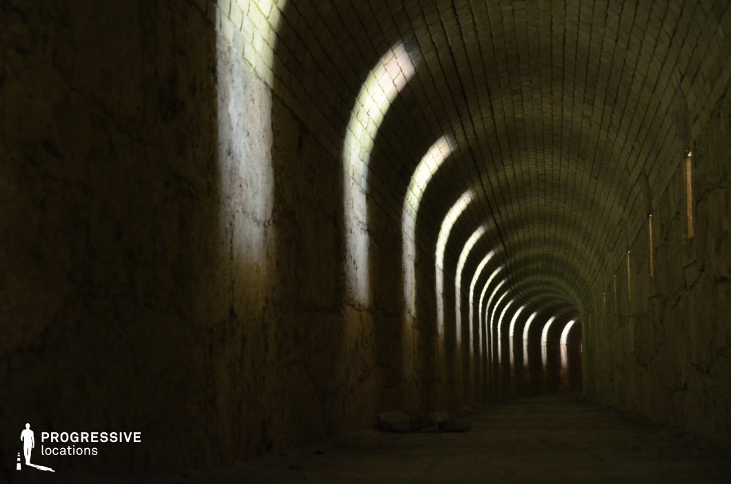 The image showcases a long, narrow tunnel with a curved brick ceiling and rough stone walls, punctuated by a series of small, evenly spaced openings that allow beams of light to cast striking arch-shaped patterns along the passage. The textured walls and aged bricks contribute to a sense of history and mystery, while the diminishing perspective draws the eye toward a distant, shadowed end. The interplay of light and darkness creates an eerie, cinematic atmosphere, making this location ideal for psychological thrillers, historical dramas, or horror films, particularly for chase sequences, hidden passageway discoveries, or suspenseful moments of entrapment.