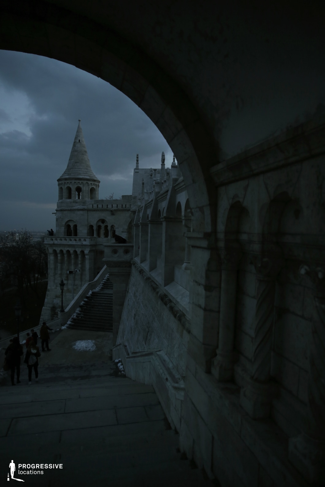 This image presents an imposing stone structure viewed from within a shadowed archway, framing a striking tower and a series of ornate colonnades. The fortress-like architecture, with its turrets, symmetrical staircases, and decorative archways, showcases a neo-Romanesque style, characterized by intricate carvings and a grand sense of scale. The steep stone steps lead toward the monumental towers, enhancing the perspective and depth. The contrast between the dark foreground and the lighter, detailed stone surfaces heightens the dramatic effect. This location is ideal for fantasy epics, historical dramas, or gothic thrillers, offering an evocative setting for royal intrigue, secret rendezvous, or heroic ascents.