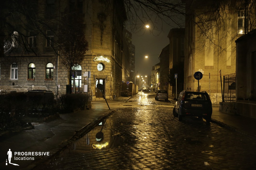 This atmospheric urban street is defined by its aged cobblestone pavement, glistening under the glow of scattered streetlights. The surrounding historic buildings, featuring arched windows and textured facades, evoke a sense of timeworn charm, while a neon-lit establishment adds a subtle focal point. Puddles on the ground create reflections that enhance the moody, noir-like aesthetic. The winding street, lined with parked vehicles and urban elements, extends into the distance, creating depth and perspective. This location is ideal for crime dramas, neo-noir thrillers, or mystery narratives, offering a visually rich and cinematic backdrop for tense encounters, covert pursuits, or contemplative late-night scenes.