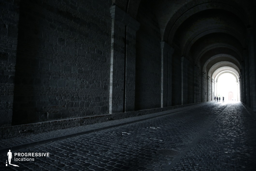 This image captures a vast, arched stone tunnel with a textured cobblestone path, leading toward a bright opening in the distance. The high, symmetrical arches create a sense of depth and scale, while the interplay of deep shadows and intense light at the tunnel’s exit enhances the dramatic contrast. The worn brick walls and rough stone surfaces emphasize the historical and almost foreboding nature of the passageway. This location is ideal for thriller, mystery, or historical films, serving as a striking backdrop for chase sequences, clandestine meetings, or symbolic journeys from darkness to revelation.