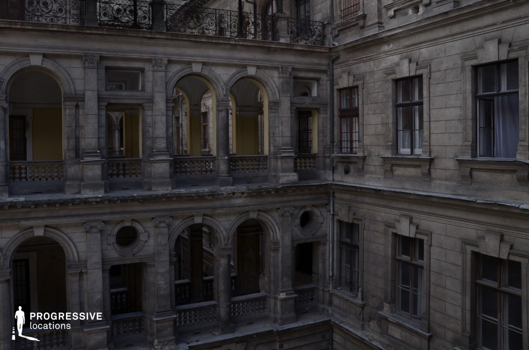 The image features a multi-story inner courtyard enclosed by a grand neoclassical stone facade with intricate detailing. The architecture is defined by large arched openings, balustraded walkways, and sculpted ornamental elements, evoking a sense of faded aristocratic elegance. The weathered stone and dimly lit passageways contribute to an aged and atmospheric quality. The symmetry of the arches and the interplay of light and shadow create a visually compelling depth. This location is ideal for historical dramas, period thrillers, or mystery films, providing an evocative setting for intrigue, clandestine meetings, or dramatic confrontations.