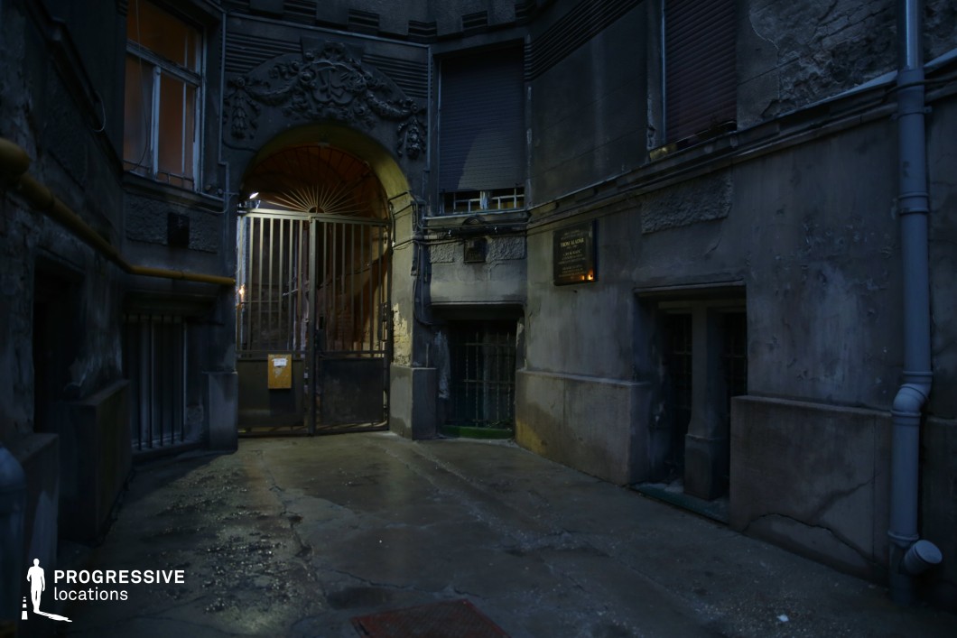 This image depicts a dimly lit, enclosed courtyard featuring an intricately carved archway with a heavy metal gate, partially ajar. The surrounding walls, marked by peeling paint, exposed wiring, and barred windows, suggest an aged and neglected structure. The wet pavement reflects the minimal light, adding to the scene’s cold, foreboding atmosphere. The blend of classical ornamentation and urban decay creates a setting that is both elegant and unsettling. This location is ideal for crime thrillers, noir films, or psychological dramas, providing a tense backdrop for secret meetings, escape sequences, or moments of eerie solitude.