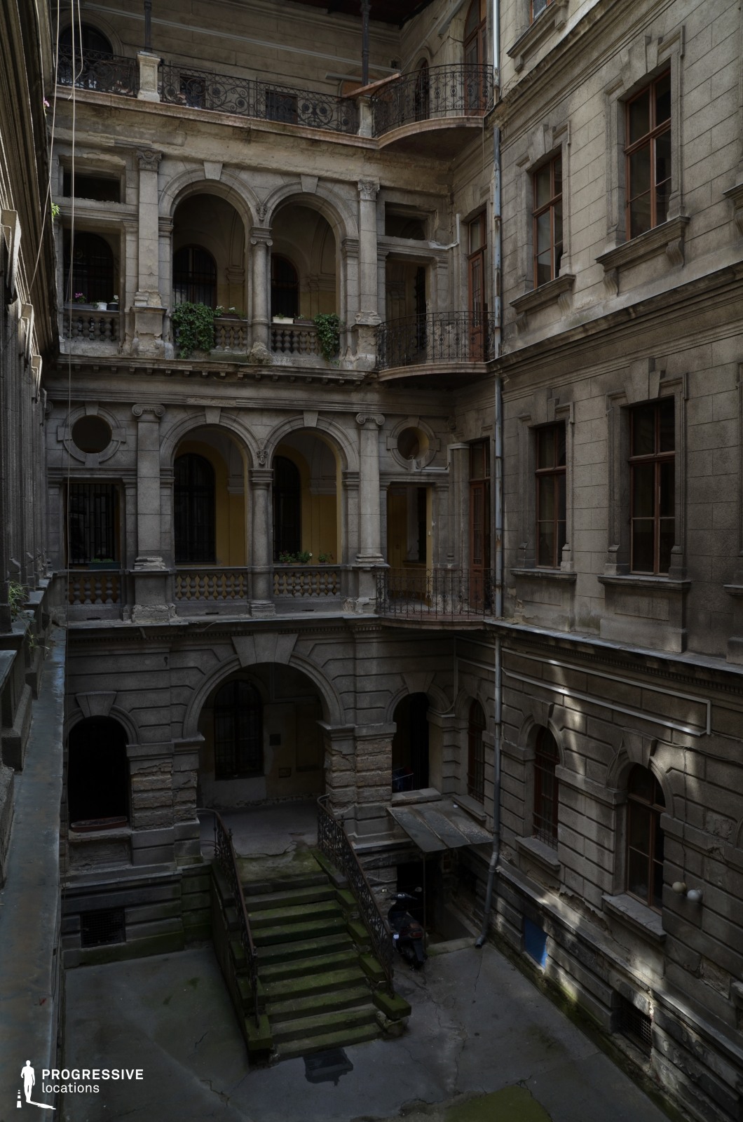 This image captures a multi-level inner courtyard of an aged historic building, featuring ornate archways, decorative balustrades, and intricate wrought-iron railings. The pale stone facade, though elegant, shows clear signs of weathering, with moss-covered steps and crumbling edges adding to the structure’s worn yet dignified charm. The layered balconies, framed by stately columns and detailed masonry, create depth and a sense of grandeur. Sunlight filtering through highlights the contrast between decay and beauty. This location is ideal for period dramas, mystery thrillers, or noir-style films, offering a setting rich with history, secrecy, and architectural splendor.