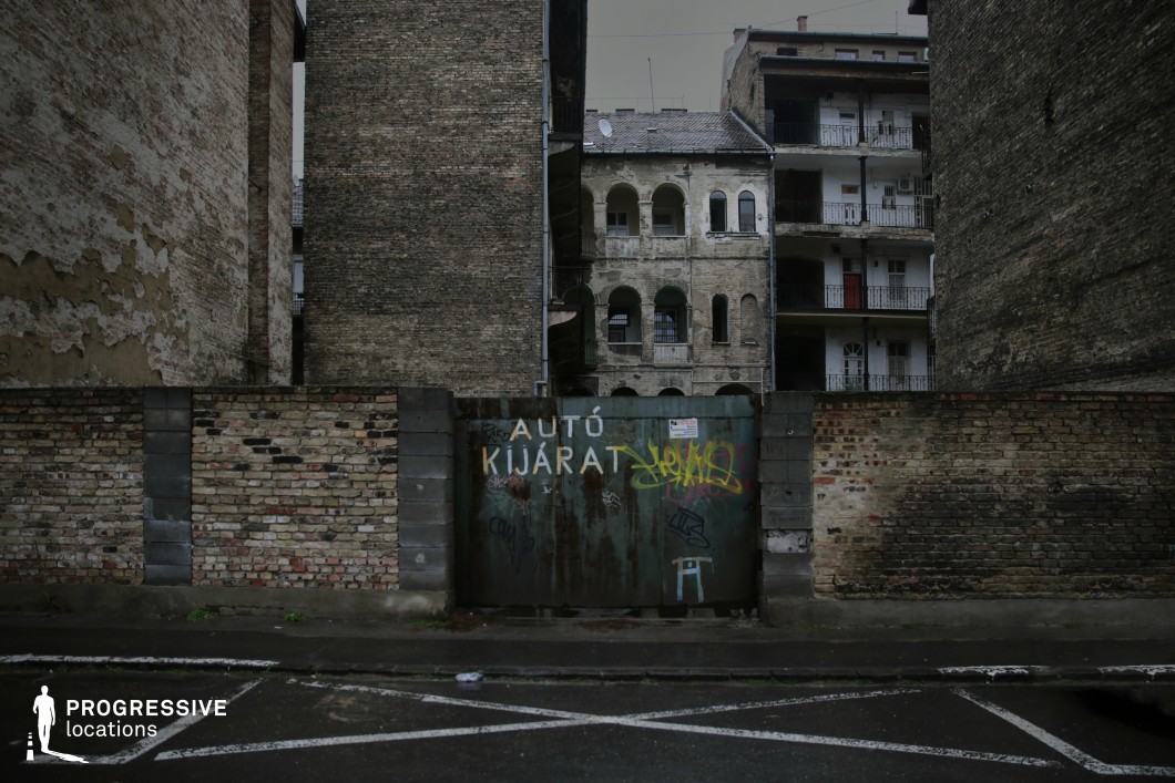 This image features an aged urban courtyard enclosed by deteriorating brick walls and tall, worn buildings with exposed balconies and arched openings. The foreground is dominated by a graffiti-covered metal gate set into the rough brickwork, adding an element of urban decay. The surrounding structures, with their peeling facades and darkened corridors, evoke a sense of abandonment and history. The muted color palette and gritty textures enhance the stark, industrial aesthetic, making this location ideal for crime dramas, dystopian thrillers, or urban noir films. It serves as a compelling backdrop for clandestine meetings, escape sequences, or narratives exploring forgotten city spaces.