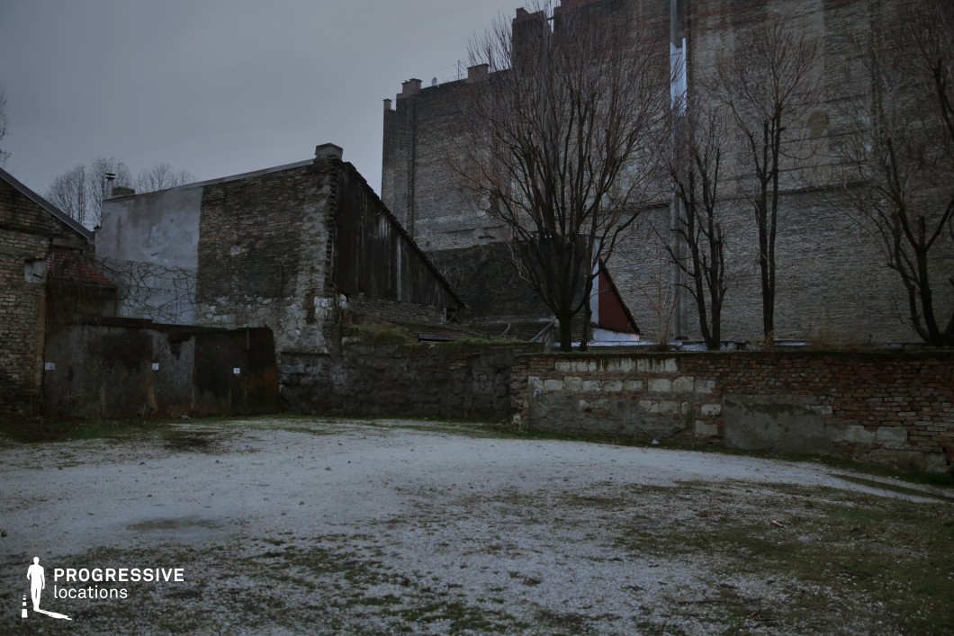 This image showcases an abandoned urban courtyard enclosed by weathered brick walls and dilapidated buildings. The uneven ground, a mix of gravel and sparse grass, adds to the stark and unkempt appearance of the space. Bare trees stand against the backdrop of towering, industrial-style brick facades, reinforcing the cold, desolate atmosphere. The mixture of old stone, patched masonry, and rusted metal elements suggests a history of industrial or residential decay. This location is ideal for dystopian thrillers, crime dramas, or post-apocalyptic settings, serving as a backdrop for tense encounters, secret exchanges, or scenes of urban decay and abandonment.