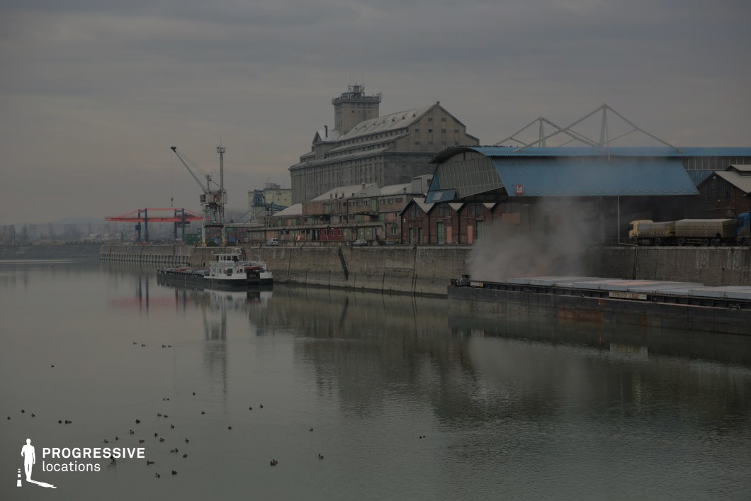 The image showcases a working industrial riverbank lined with large warehouses, cranes, and a massive concrete embankment. A barge docked at the quay releases steam or smoke, adding a sense of activity to the otherwise quiet setting. The architecture blends utilitarian metal-roofed structures with old brick warehouses, while a towering grain silo dominates the skyline, emphasizing the site's functional and historical significance. The still water reflects the muted industrial tones, contributing to a moody and atmospheric backdrop. This location is ideal for crime dramas, neo-noir thrillers, or espionage films, providing a gritty, urban waterfront setting for smuggling operations, clandestine meetings, or tense pursuit sequences.